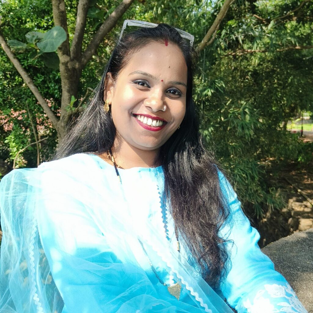 Woman smiling, wearing a blue kurta sitting in front of a foliage background